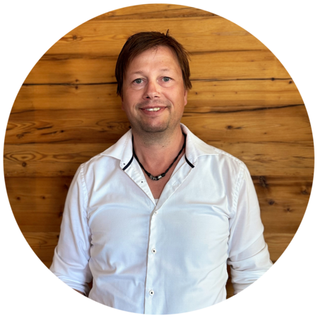 Mann mit hellbraunen, kurzen Haaren und einem Dreitagebart, trägt ein weißes Hemd und eine schwarze Halskette.